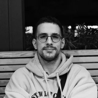 Pavle Tošić, crno-bijeli portret: kratka kosa, okrugle naočare i brada, sjedi na drvenoj klupi.