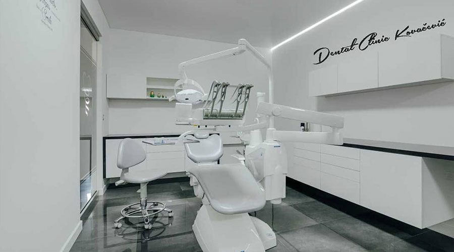 Bijela ordinacija sa stomatološkom stolicom i radnom lampom; na zidu iznad pultova rukom ispisano ime Dental Clinic Kovačević