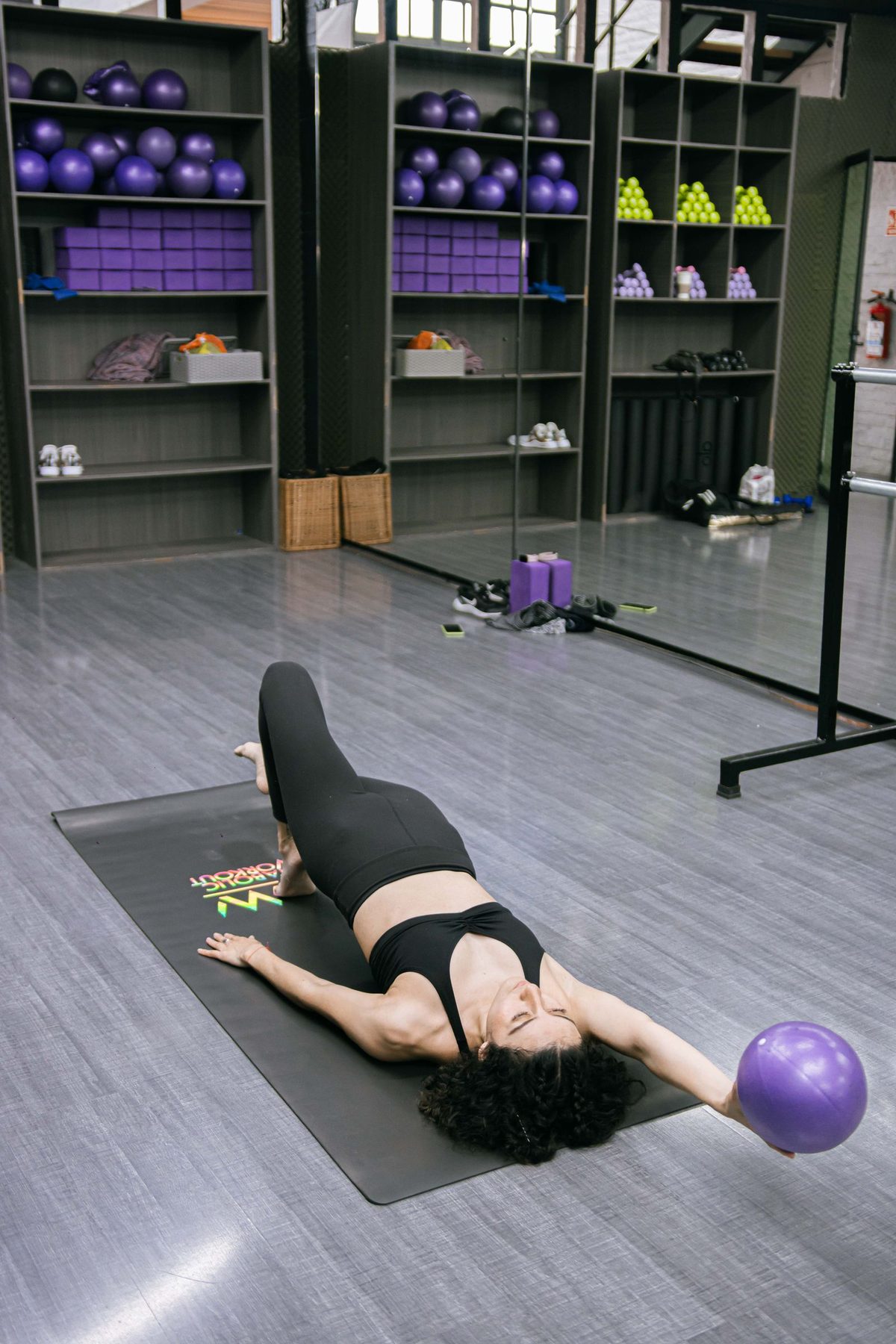 Vježbačica leži na strunjači i vodi malu pilates loptu u ispruženoj ruci, koljena savijena; iza nje police sa loptama, blokovima i balet šipka