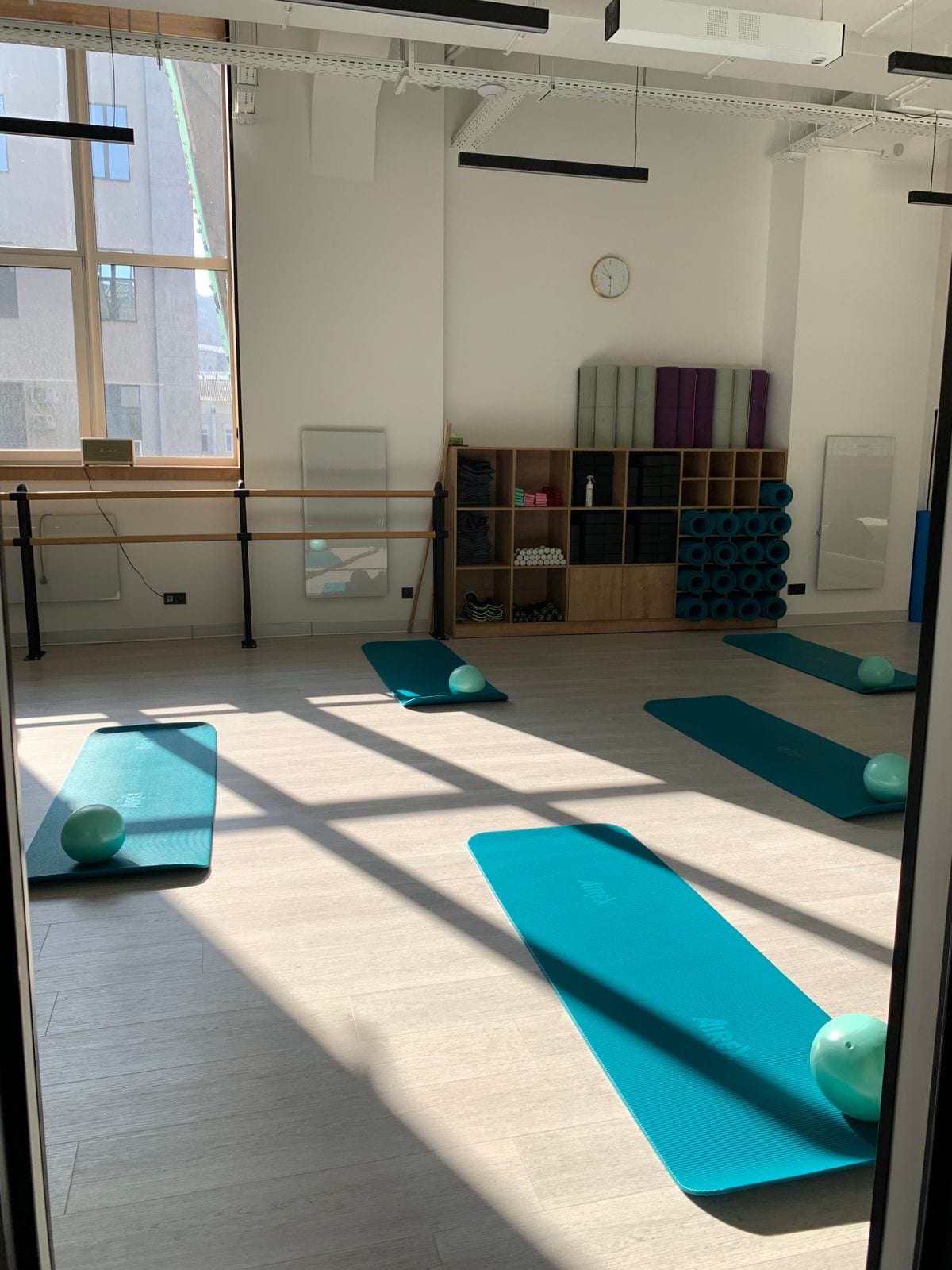 Prazna sala za mat pilates: strunjače sa malim loptama poređane po drvenom podu, balet šipka uz zid i polica sa smotanim strunjačama i rekvizitima, u svjetlu sa velikog prozora